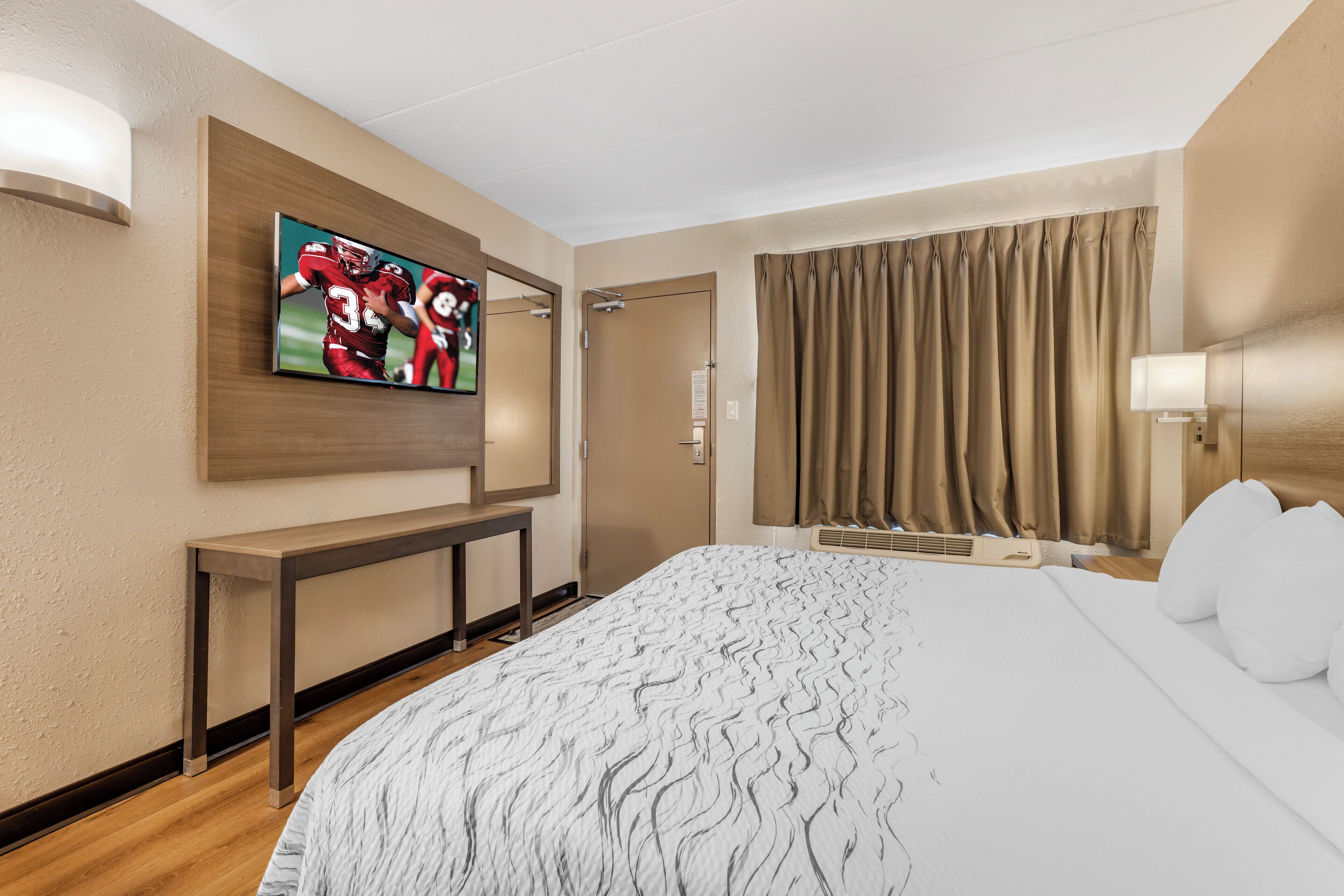 Une chambre d’hôtel dotée d’un grand lit, d’un téléviseur à écran plat fixé au mur, d’une table de chevet, d’une fenêtre avec des rideaux et d’un climatiseur mural.