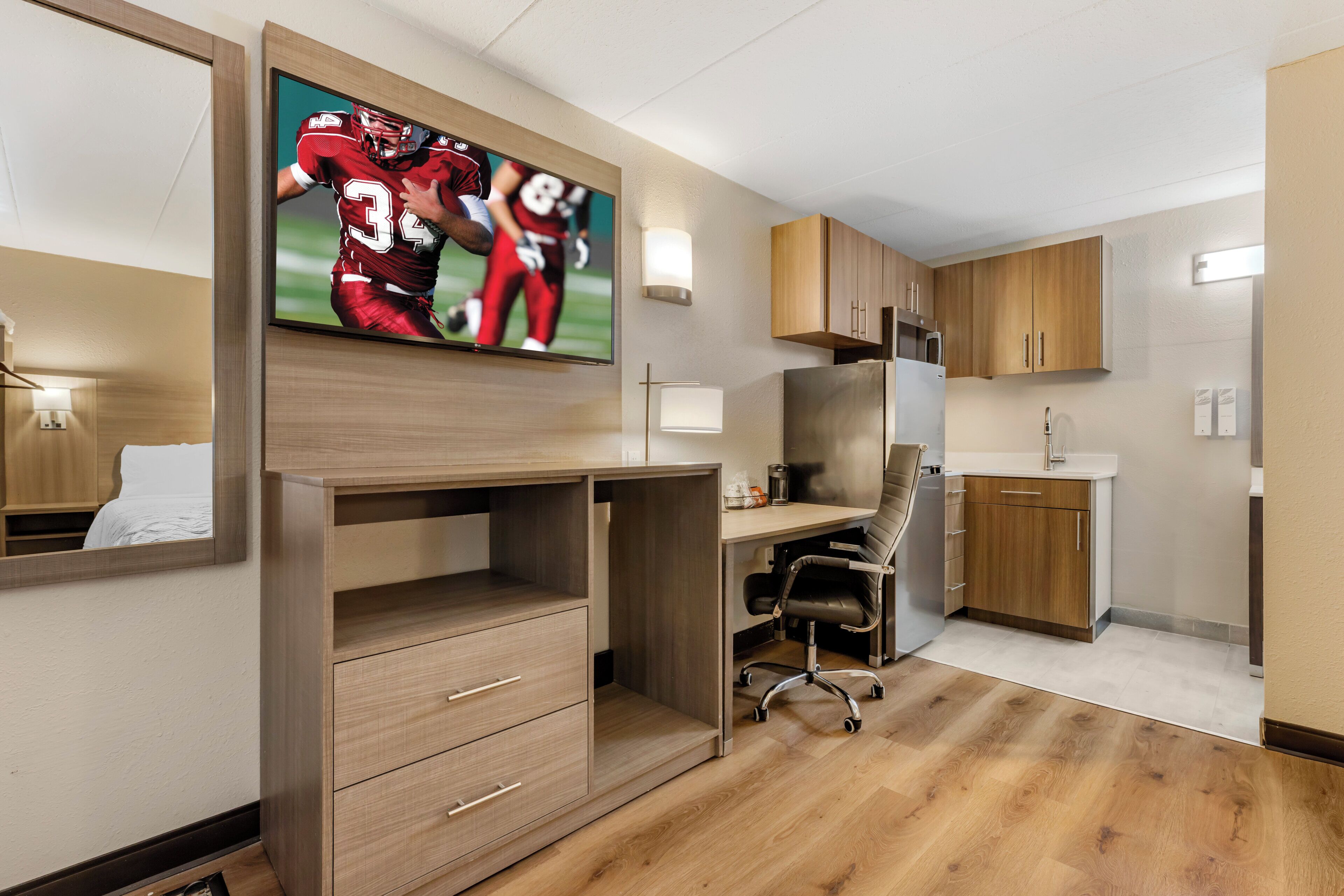 A hotel room with a wooden desk, a chair, a flat-screen TV mounted on the wall, a kitchen area with wooden cabinets, and a refrigerator.