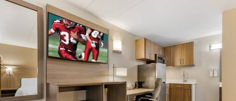 A hotel room with a wooden desk, a chair, a flat-screen TV mounted on the wall, a kitchen area with wooden cabinets, and a refrigerator.