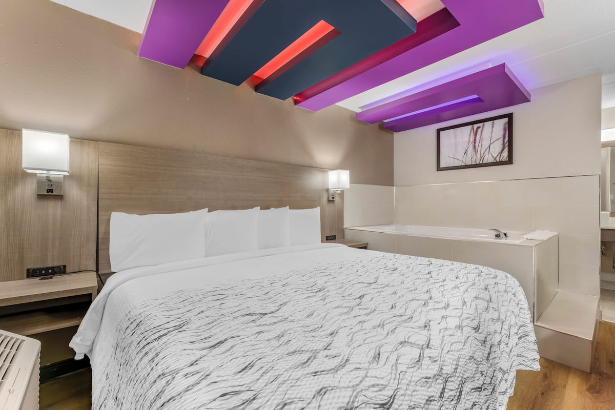 A modern hotel room with a bed, bedside tables, a nightstand, a wall-mounted light, and a ceiling with colorful lighting.