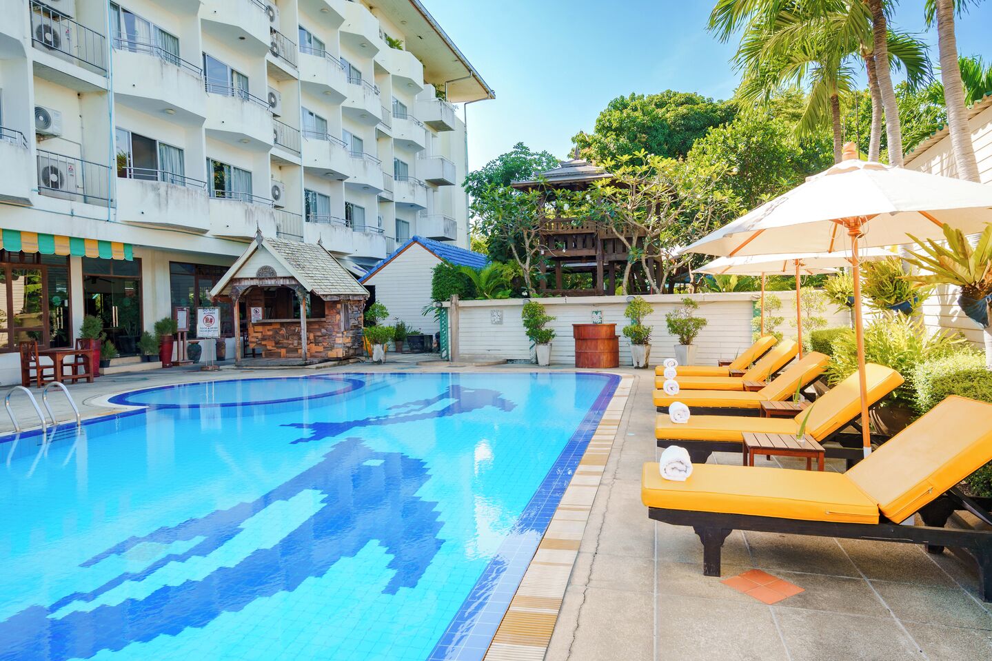 A swimming pool with lounge chairs and umbrellas, surrounded by a building with balconies and a small structure with a tiled roof.