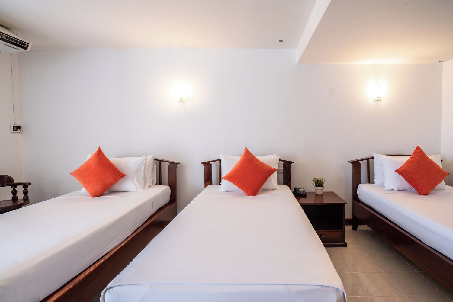 Habitación de hotel con tres camas individuales, cada una con ropa de cama blanca y almohadas naranjas. Hay una mesita de noche pequeña con una planta y un teléfono encima. El cuarto tiene un diseño sencillo y limpio, con paredes blancas y un cabecero de madera en cada cama.