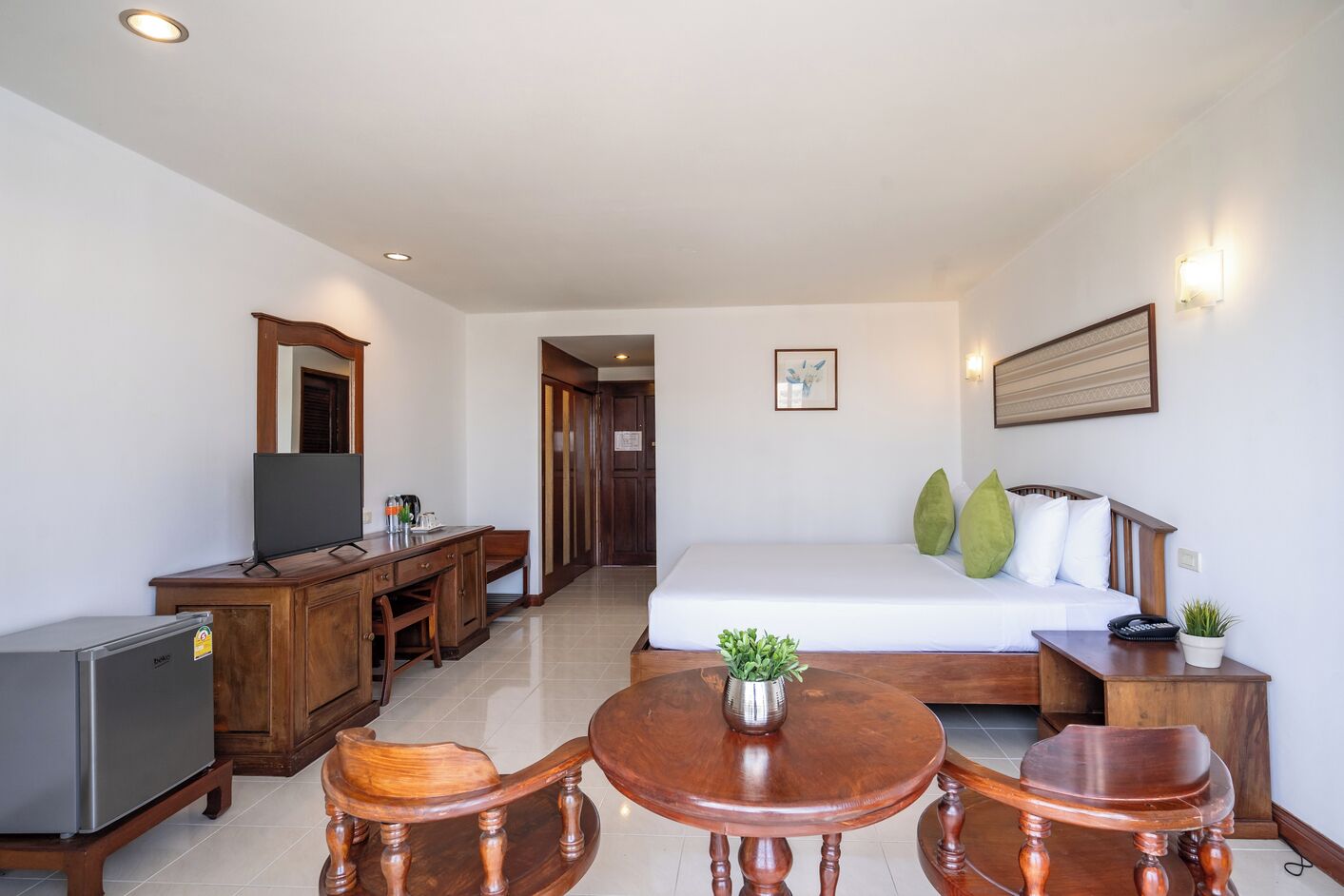A hotel room with a bed, a desk with a TV, a dining table, and a small plant.