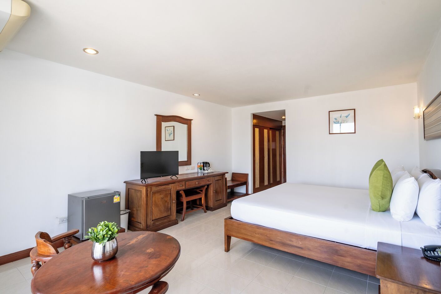 A hotel room with a wooden desk, a television, a bed with white bedding, a small table with a plant, and a wall-mounted picture.