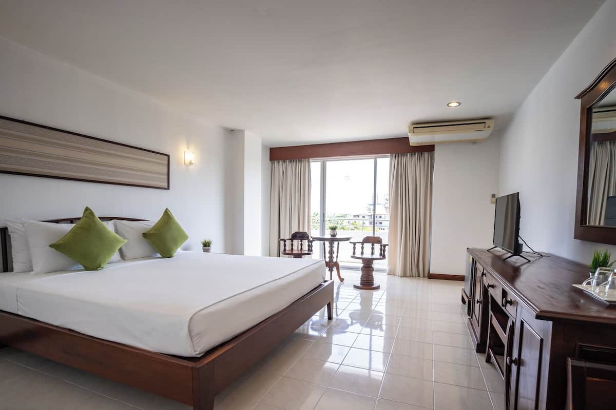 Habitación de hotel con una cama grande, un escritorio con televisor, una pequeña mesa con sillas y un balcón con cortinas.