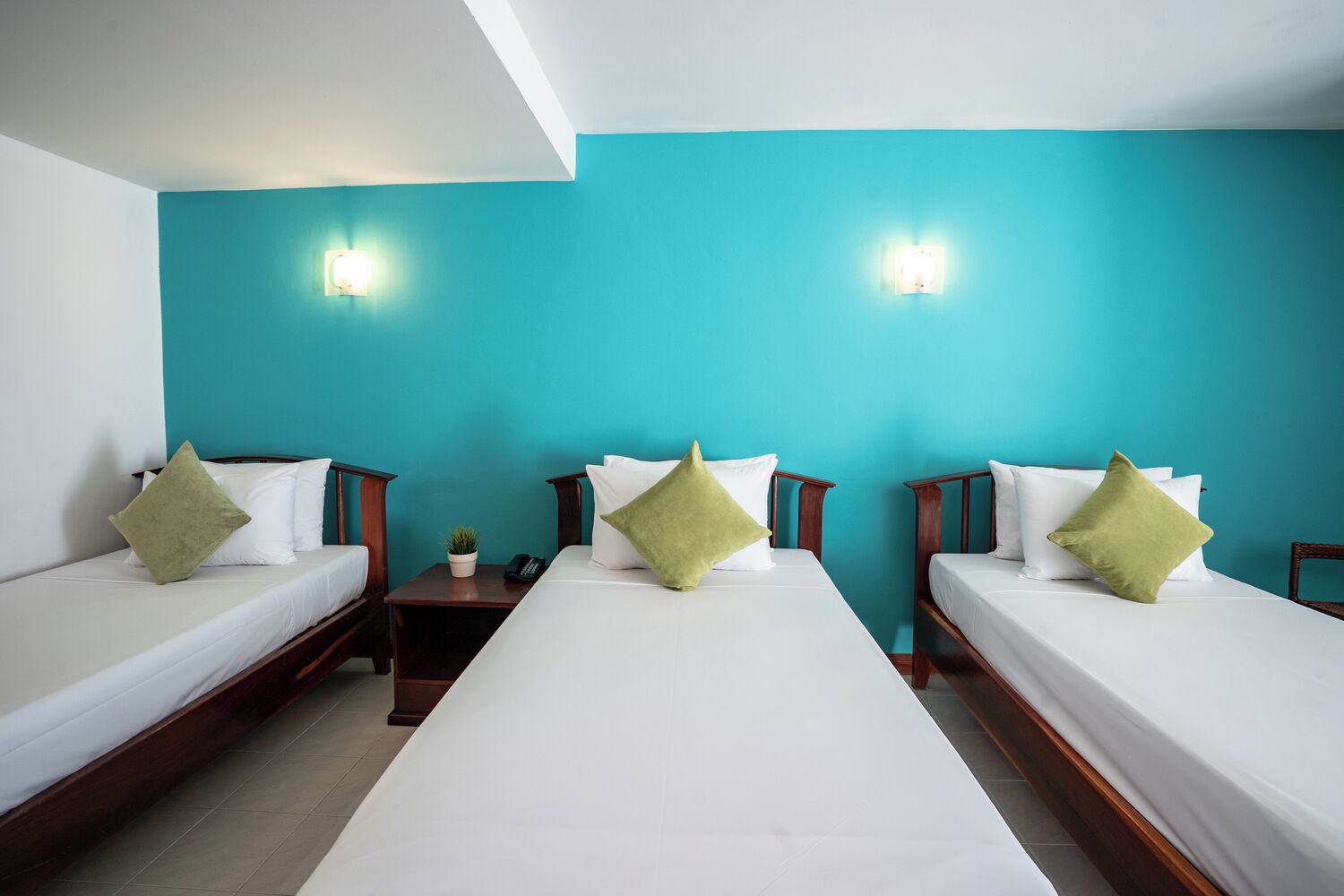 A hotel room with three single beds, each with white bedding and green pillows. There are two wall-mounted light fixtures and a small bedside table with a plant and a phone.