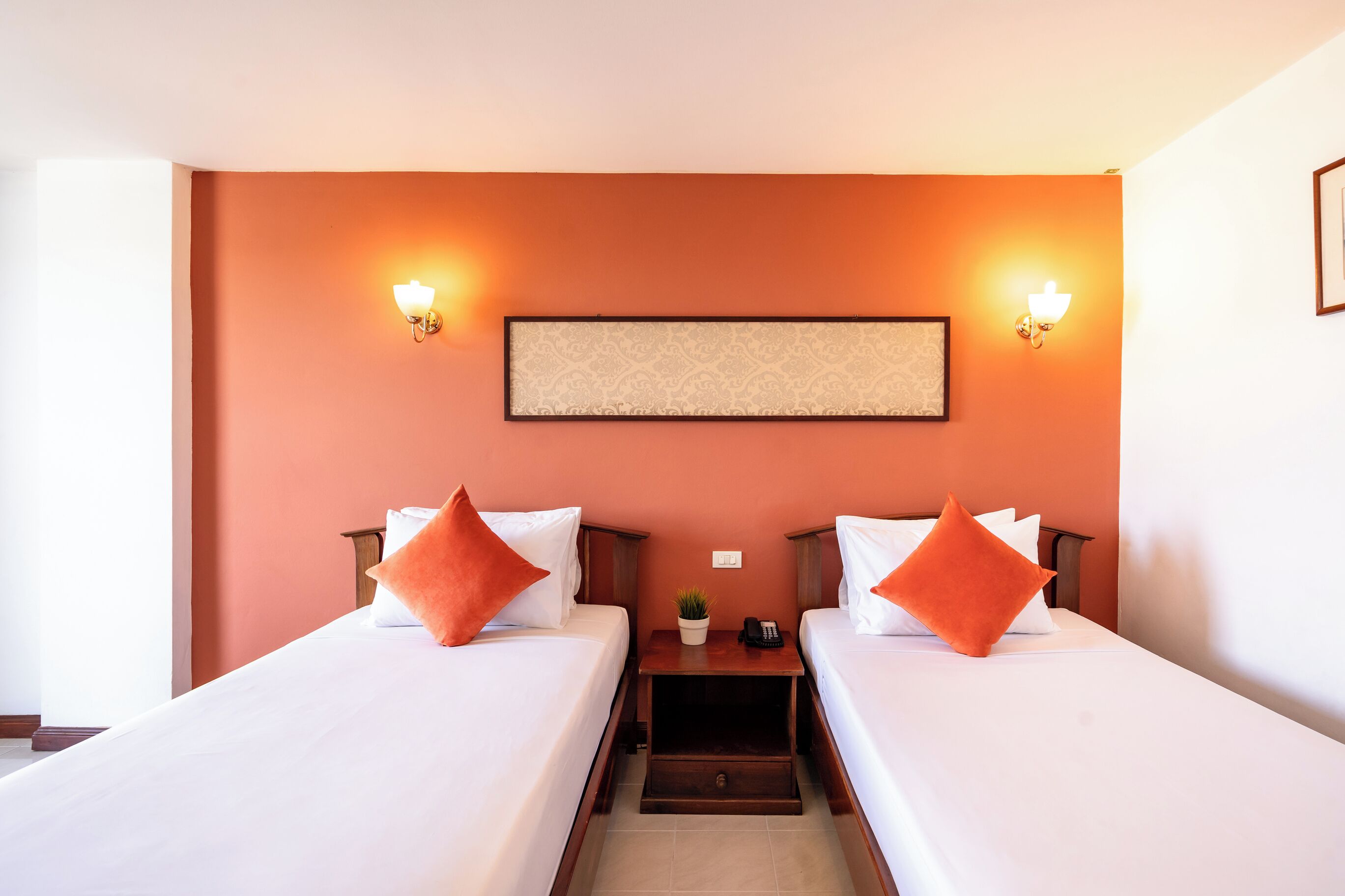 Habitación de hotel con dos camas individuales, cada una con ropa blanca y almohadas naranjas. Hay luminarias fijadas a la pared a ambos lados de una obra de arte enmarcada.