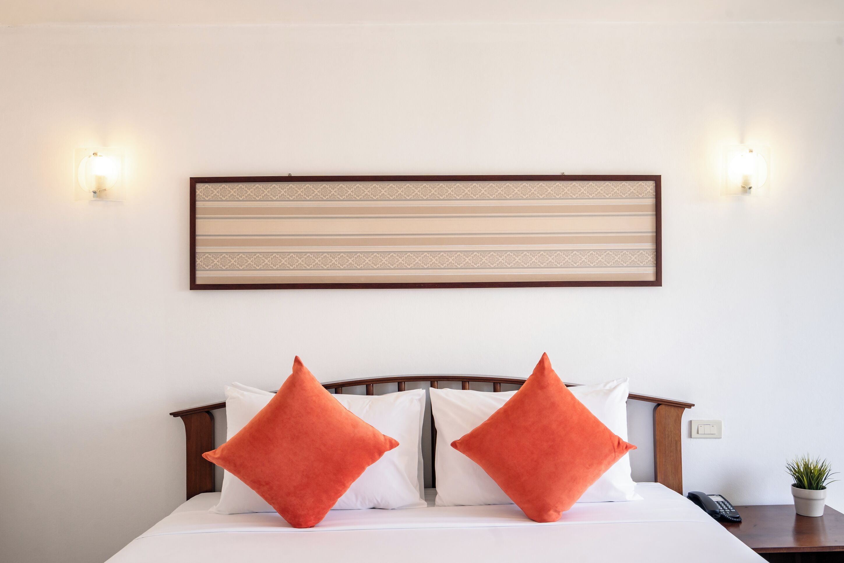 Una cama bien hecha con almohadas blancas y anaranjadas, un cabecero de madera y una lámpara fijada a la pared.