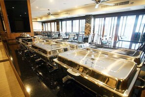 A buffet setup with multiple serving stations, a ceiling fan, and large windows allowing natural light. - Honey Inn (Pattaya)