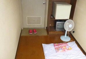 A room with a wooden floor, a white bed with a floral quilt, a standing fan, a television on a wooden shelf, and a door with a vent.