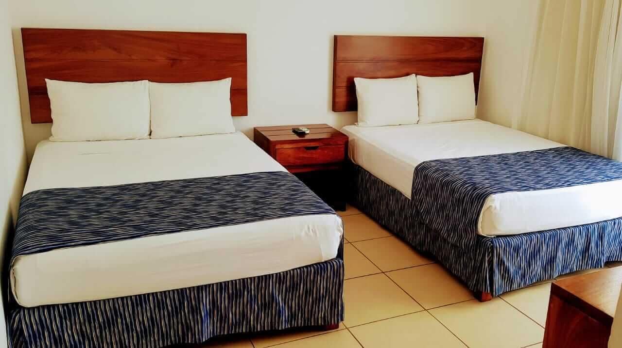 A hotel room with two beds, each with a wooden headboard, white bedding, and striped bedspreads. There is a wooden nightstand between the beds.