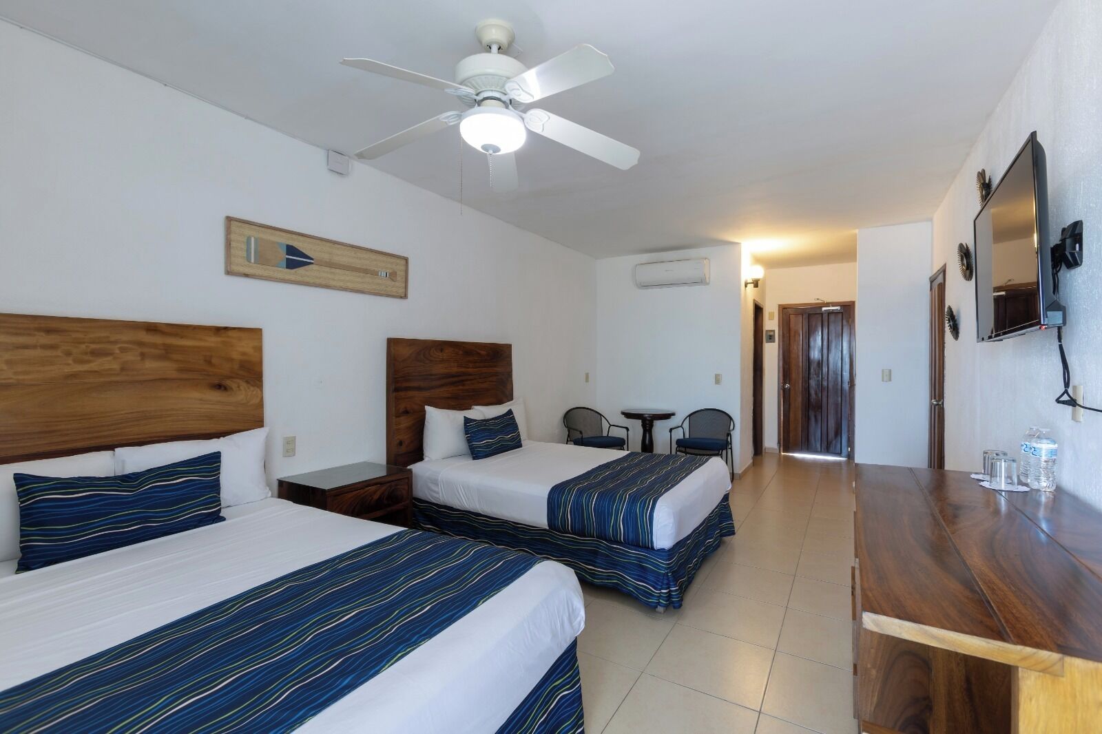 Una habitación de hotel con dos camas, un cabecero de madera, un ventilador de techo, una mesita y un televisor montado en la pared.