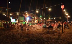 An outdoor event with tables and chairs set up on a sandy area, illuminated by string lights.
