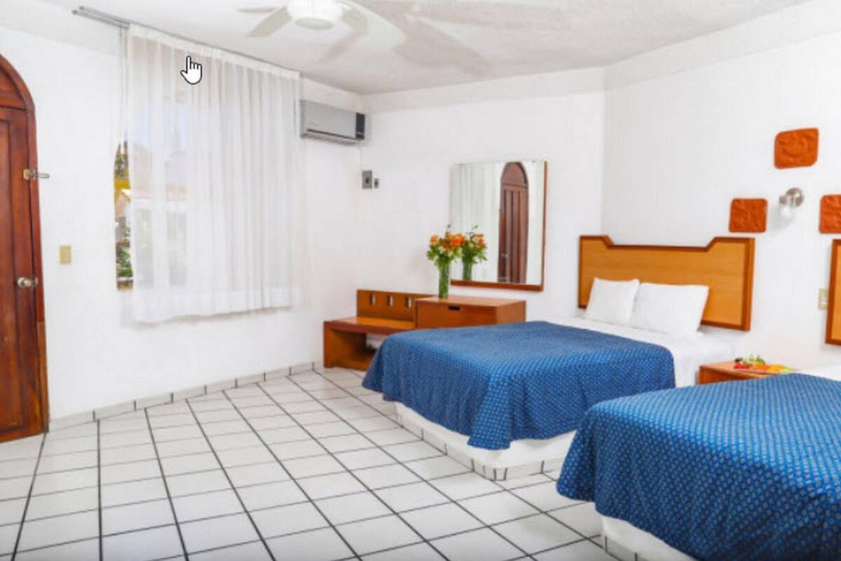 A hotel room with two beds, a wooden headboard, a bedside table, a mirror, and a window with curtains.