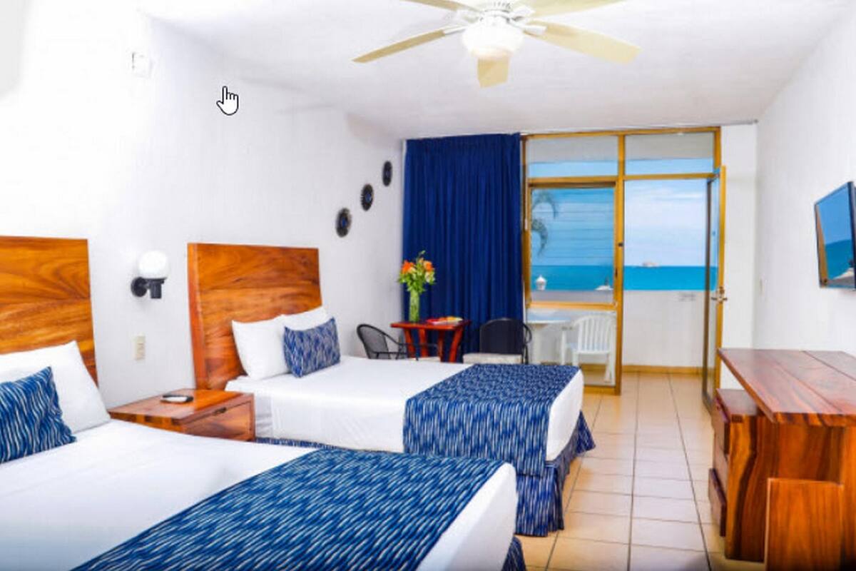 A hotel room with two beds, a desk, a chair, a small table, a ceiling fan, and a view of the ocean.