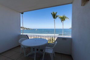 A balcony with a round table and two chairs, overlooking a beach with palm trees and the ocean.