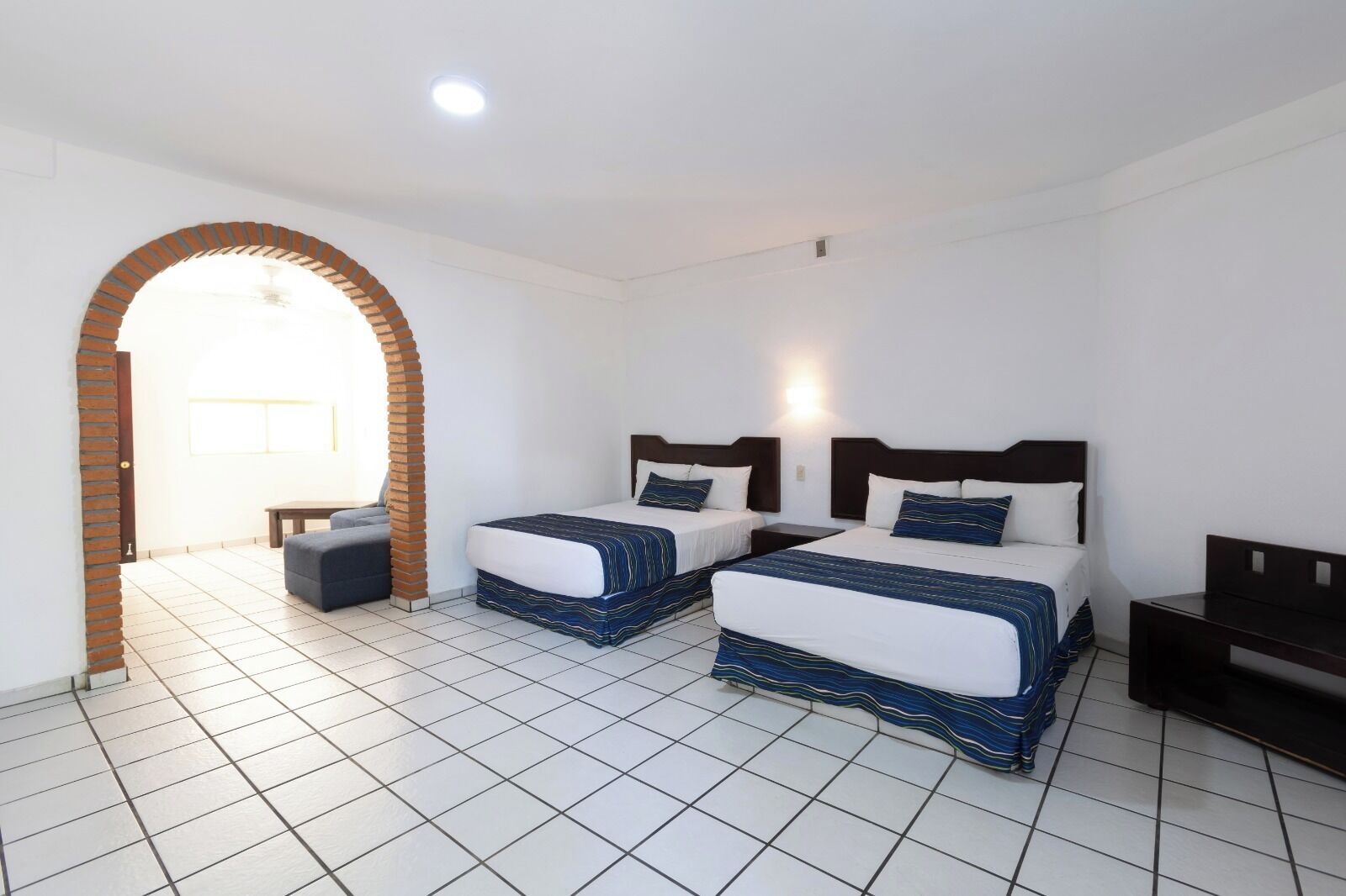 Una habitación de hotel con dos camas, una mesita, un banco y suelo de baldosas.