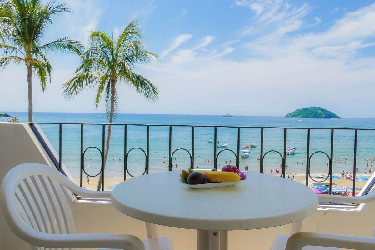 A balcony with a table and two chairs, overlooking a beach with palm trees and a clear sky.