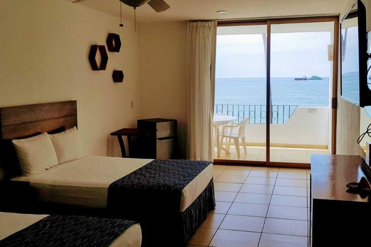 A hotel room with two beds, a small desk, a chair, and a balcony with a view of the sea.