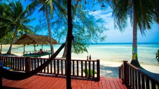 Eine Holzterrasse mit einer Hängematte, mit Blick auf einen Strand mit Palmen und einer ummauerten Hütte.