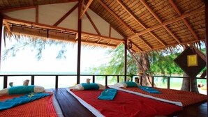 Ein Zimmer mit zwei Betten, jeweils mit roter Bettdecke und blauen Kissen, unter einem reetgedeckten Dach mit Blick auf das Meer.