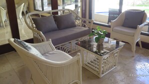 Ein Zimmer mit Korbmöbeln, darunter ein Sofa und Sessel, ein Glastisch und Blick durch große Fenster.