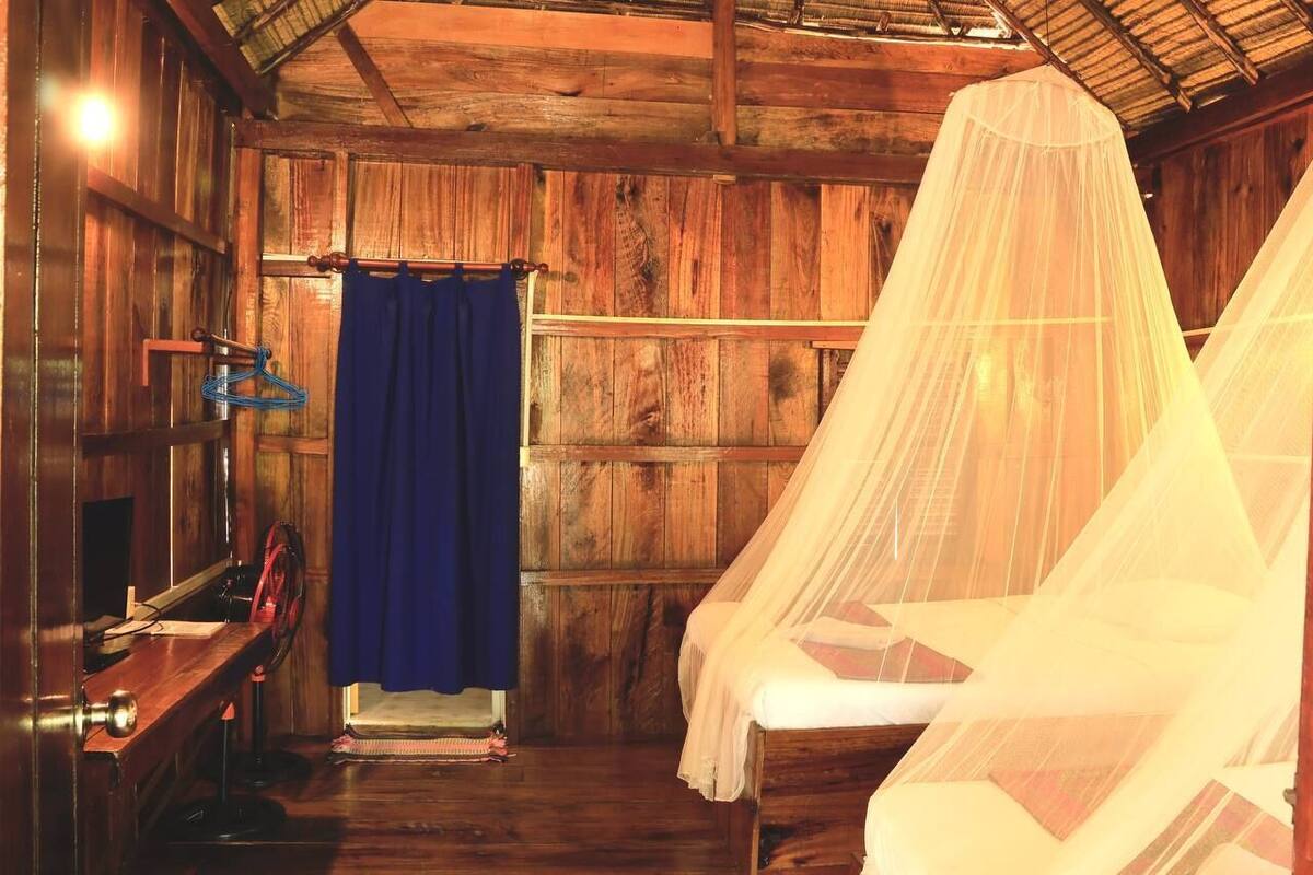 A rustic wooden room with a bed under a mosquito net, a blue curtain, and a wooden desk with a chair.