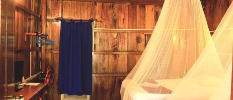Una habitación rústica de madera con una cama bajo una mosquitera, una cortina azul y un escritorio de madera con una silla.