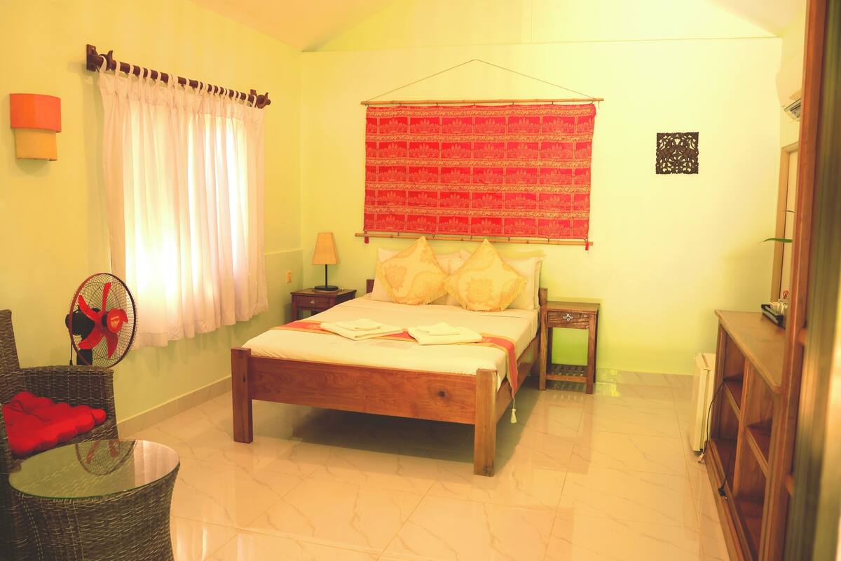 A bedroom with a wooden bed, a bedside table, a lamp, a fan, and a red curtain.