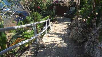 A stone staircase leading to a thatched-roof structure, surrounded by lush greenery and a clear view of the sea.