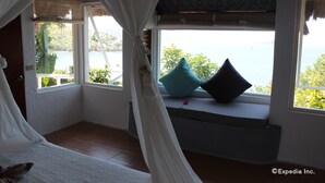 A bedroom with a bed, a window with a view of the sea, and a window seat with cushions. - Al Faro Cosmio Hotel (Busuanga)