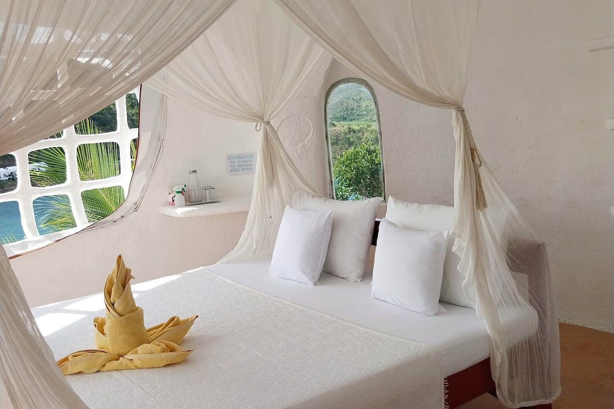 Una habitación con cama cubierta por un dosel, una ventana con vistas a vegetación y un estante con objetos.