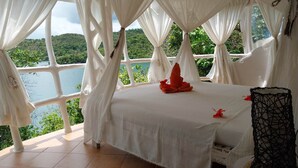 A room with a bed, white curtains, and a view of a lake and greenery.