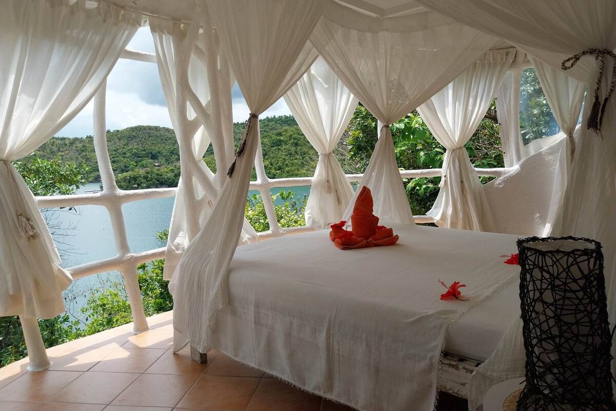 Una habitación con cama, cortinas blancas y vistas a un lago y vegetación.
