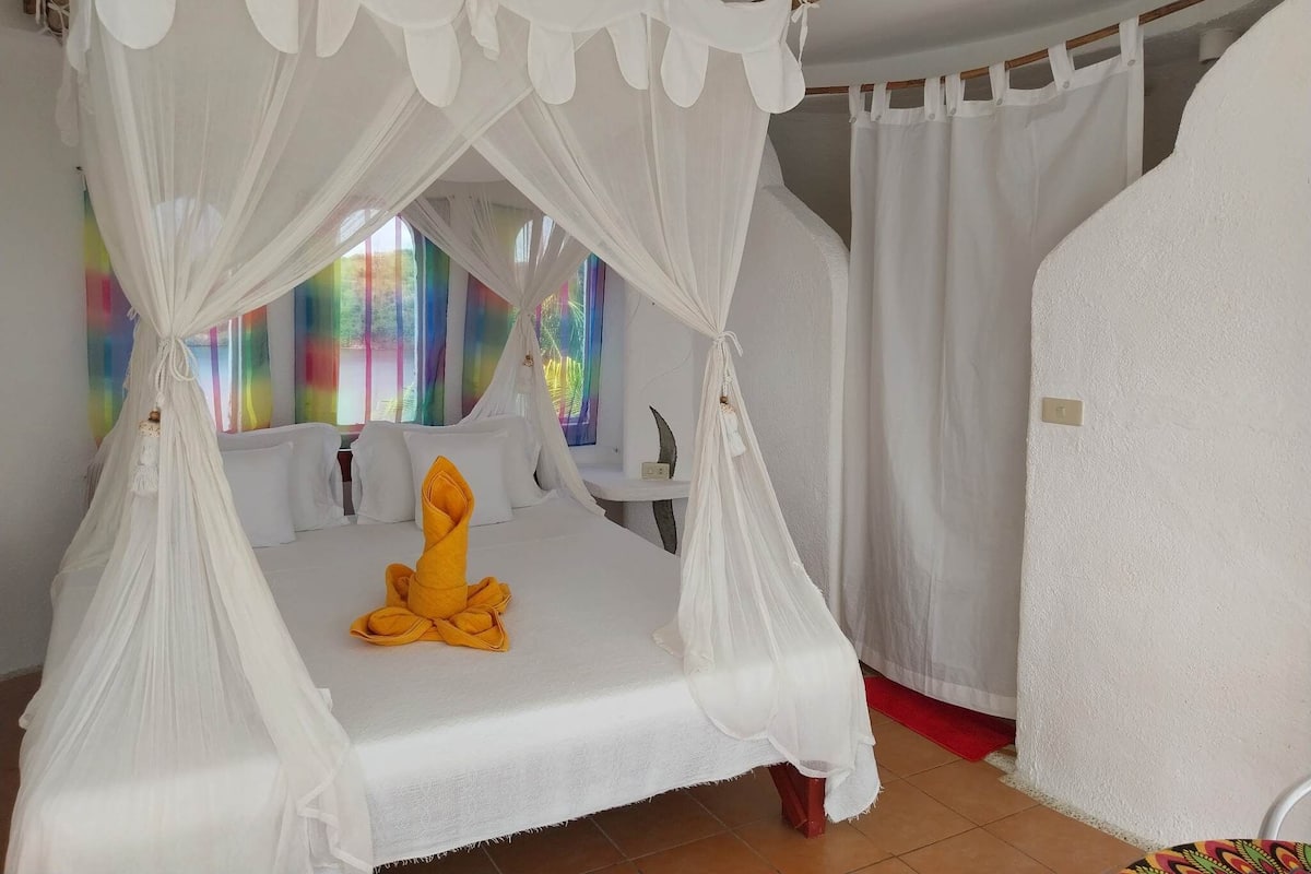 Una cama con dosel y cortinas blancas, una toalla amarilla y una vista colorida por la ventana.