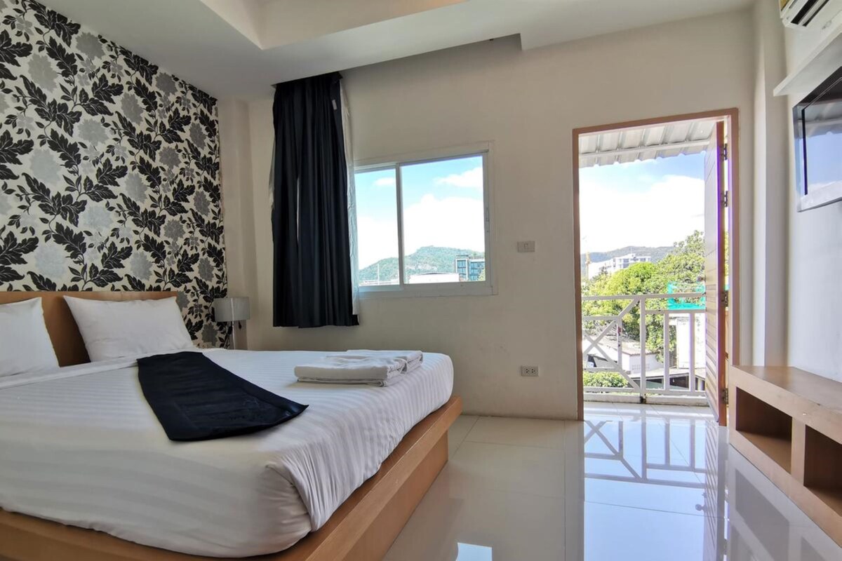 A modern hotel room with a large bed, a window with curtains, and a balcony with a view of the city.