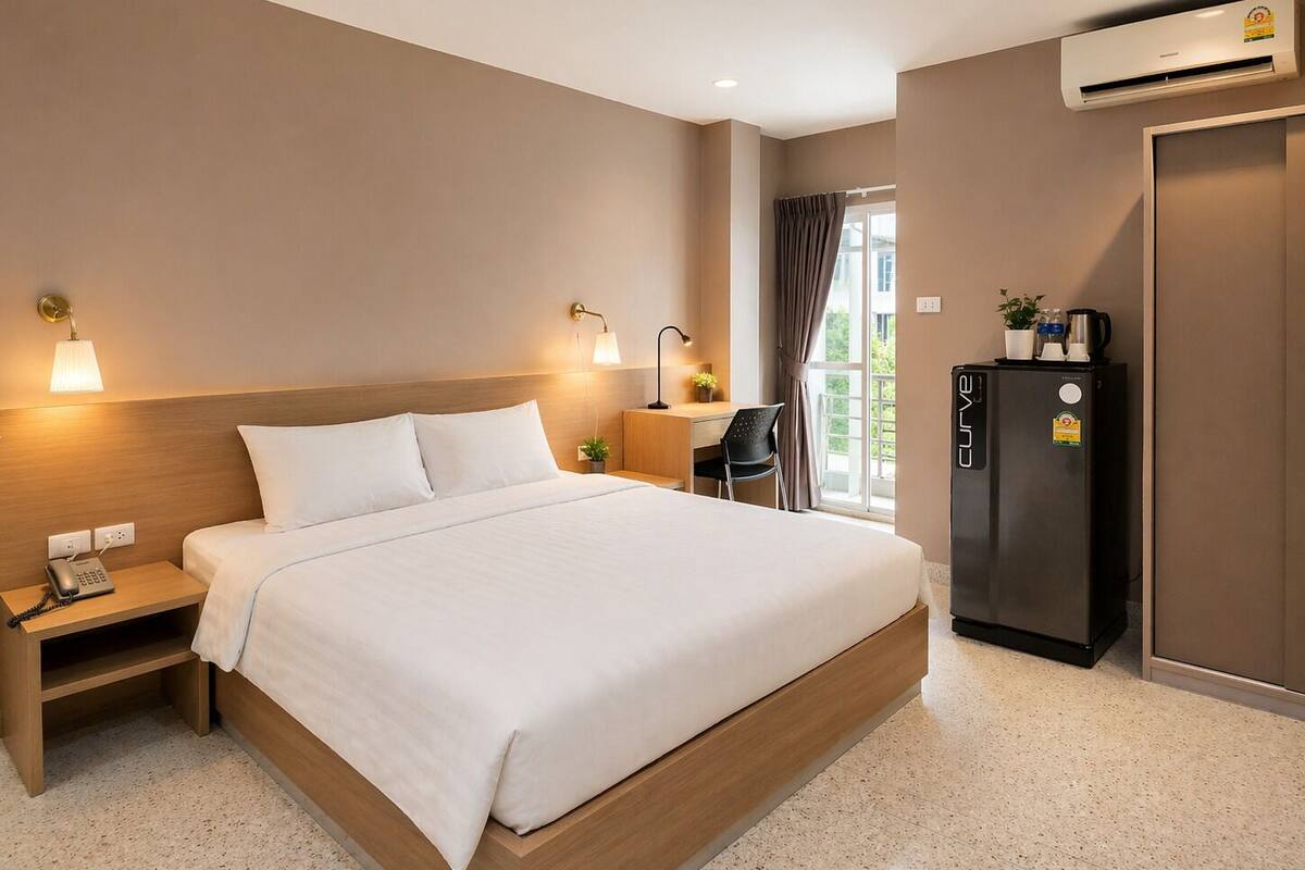 A hotel room with a bed, bedside table, lamp, desk, chair, refrigerator, and an air conditioning unit.