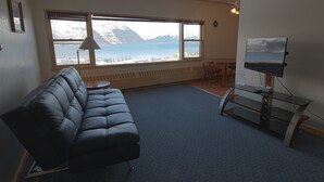 A room with a large window overlooking a body of water and mountains, a black leather couch, a glass coffee table, and a TV on a stand. - Glacier View Condo Suites (Whittier)