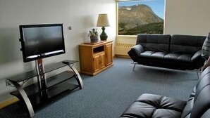 A living room with a flat-screen TV, a wooden cabinet, a black leather sofa, and a view of a mountain through a window. - Glacier View Condo Suites (Whittier)