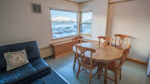 A room with a dining table and chairs, a black leather sofa, and a view of a snowy landscape through large windows. - Glacier View Condo Suites (Whittier)