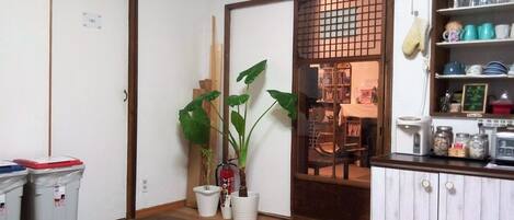 Cozinha compacta com armários de madeira, uma mesa de jantar e uma pequena geladeira. Há plantas em vasos e um ventilador na parede.