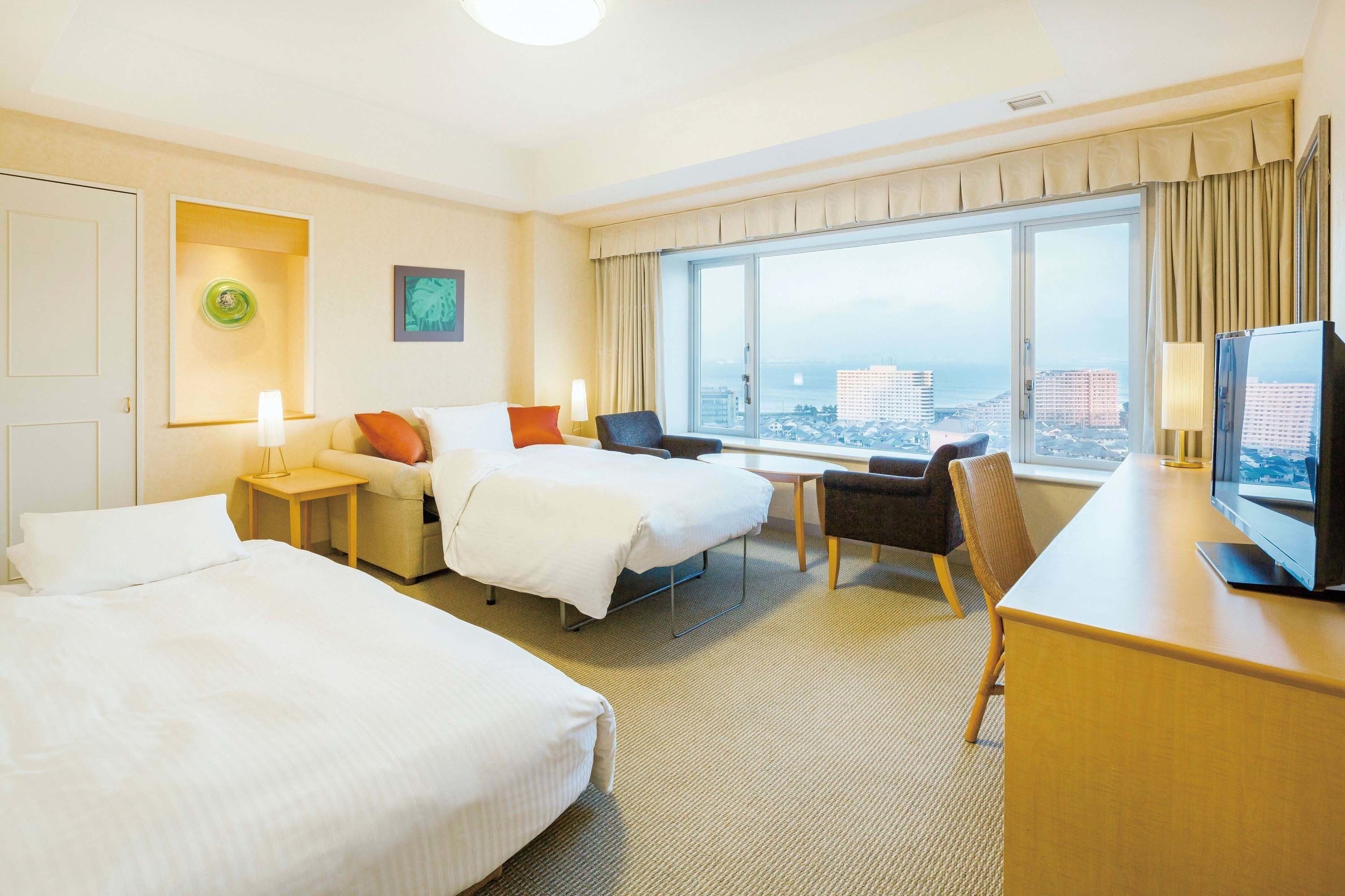 Ein Hotelzimmer mit zwei Betten, einem Schreibtisch, einem Stuhl, einem Fernseher und einem großen Fenster mit Blick auf die Stadt.