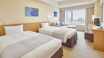 A hotel room with two beds, a desk with a TV, a small table, and a window with a view of buildings.