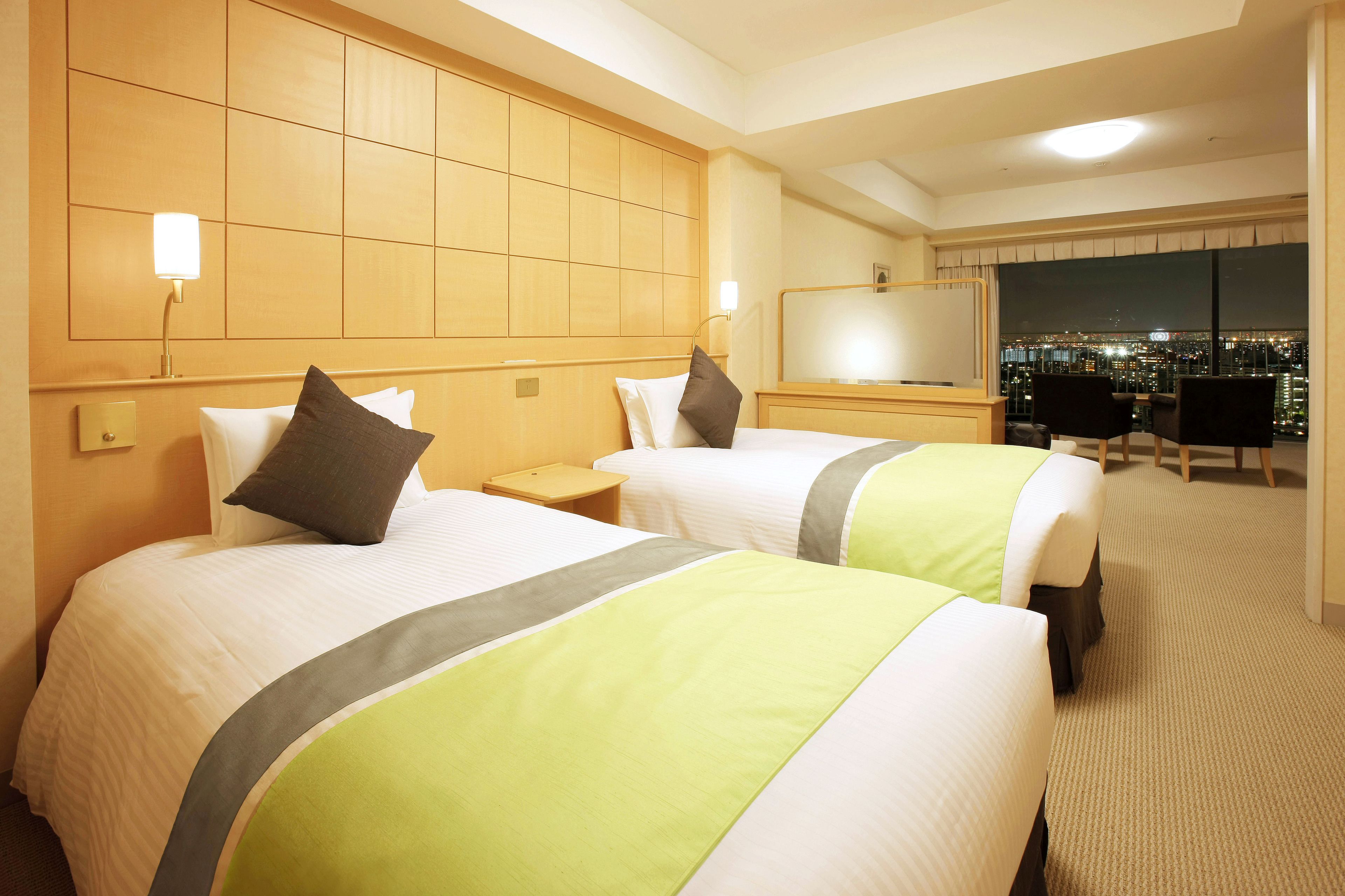 A hotel room with two beds, a nightstand, a desk, and a large window with a city view.