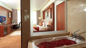 A hotel bathroom with a large mirror, a bathtub filled with rose petals, and a visible bedroom with a bed and a desk. - Denissons (Hubli)