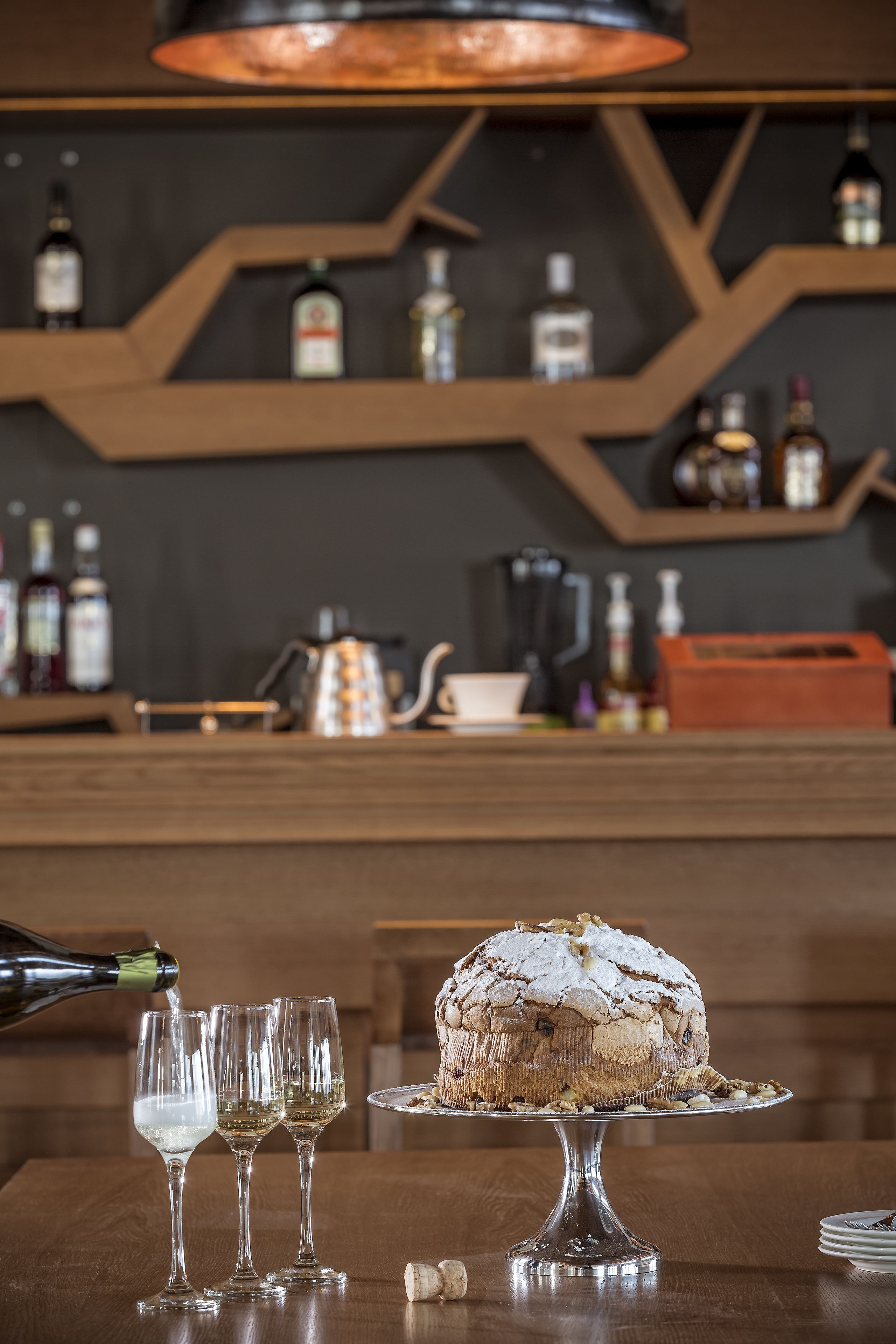 A bar area with a cake on a glass stand, three glasses of white wine, and a bottle of champagne being poured.