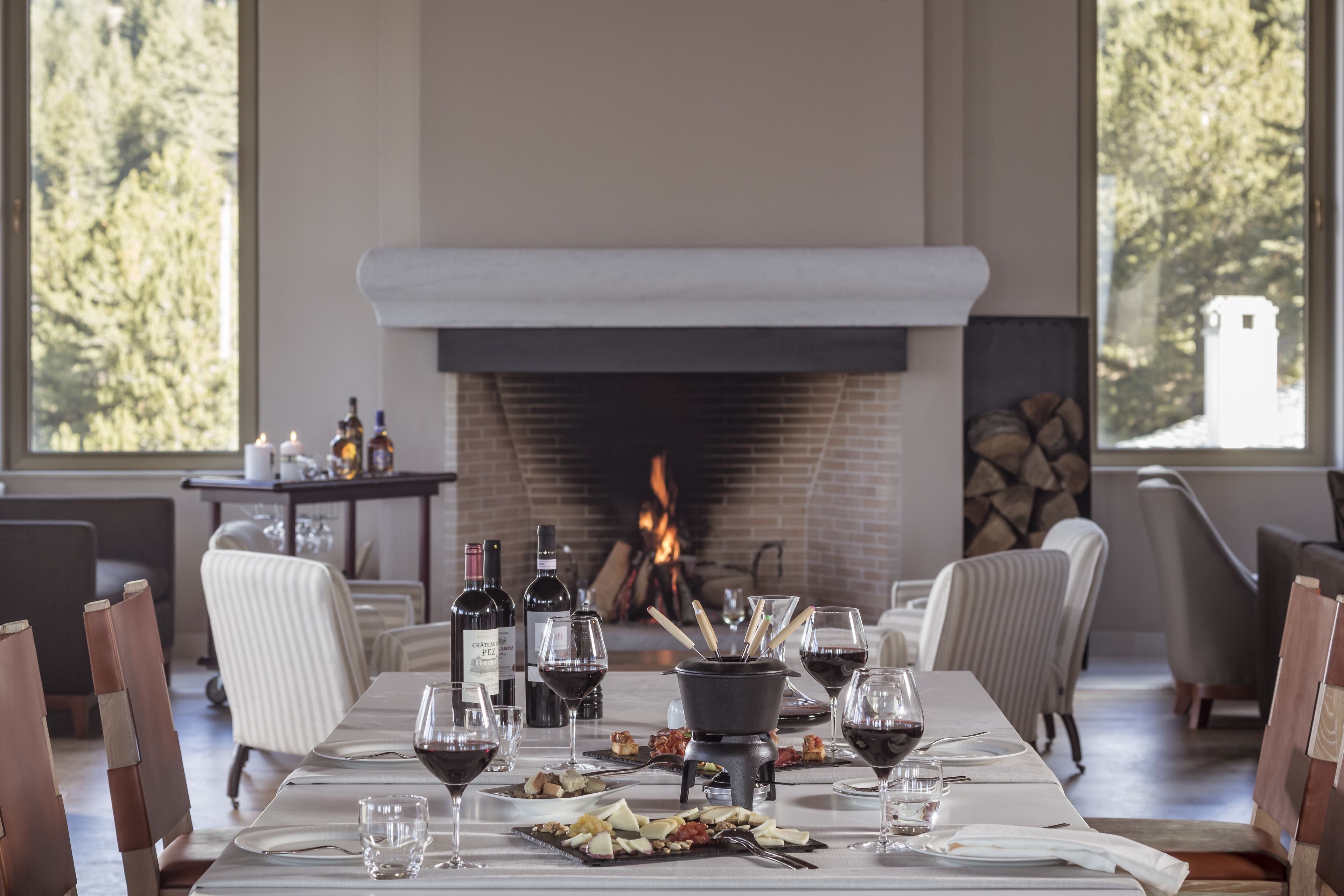 A dining area with a fireplace, wooden chairs, a long table set for a meal, and wine bottles.