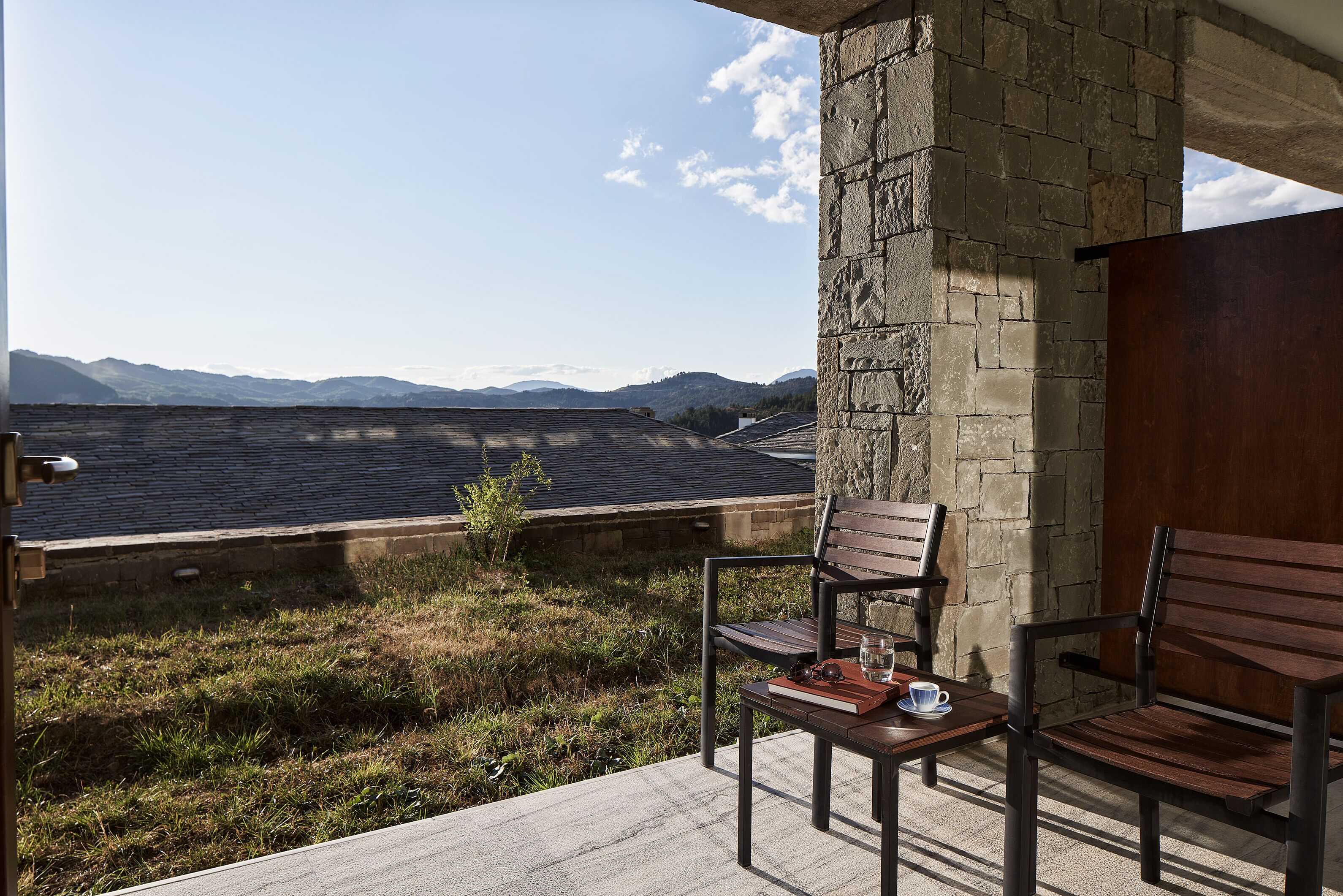 A balcony with a view of mountains, a stone wall, and two wooden chairs with a table and a book.