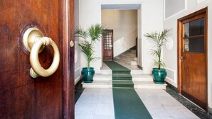 Une porte en bois avec une poignée en laiton, un sol en marbre, un escalier recouvert d’une moquette verte et deux plantes en pot.