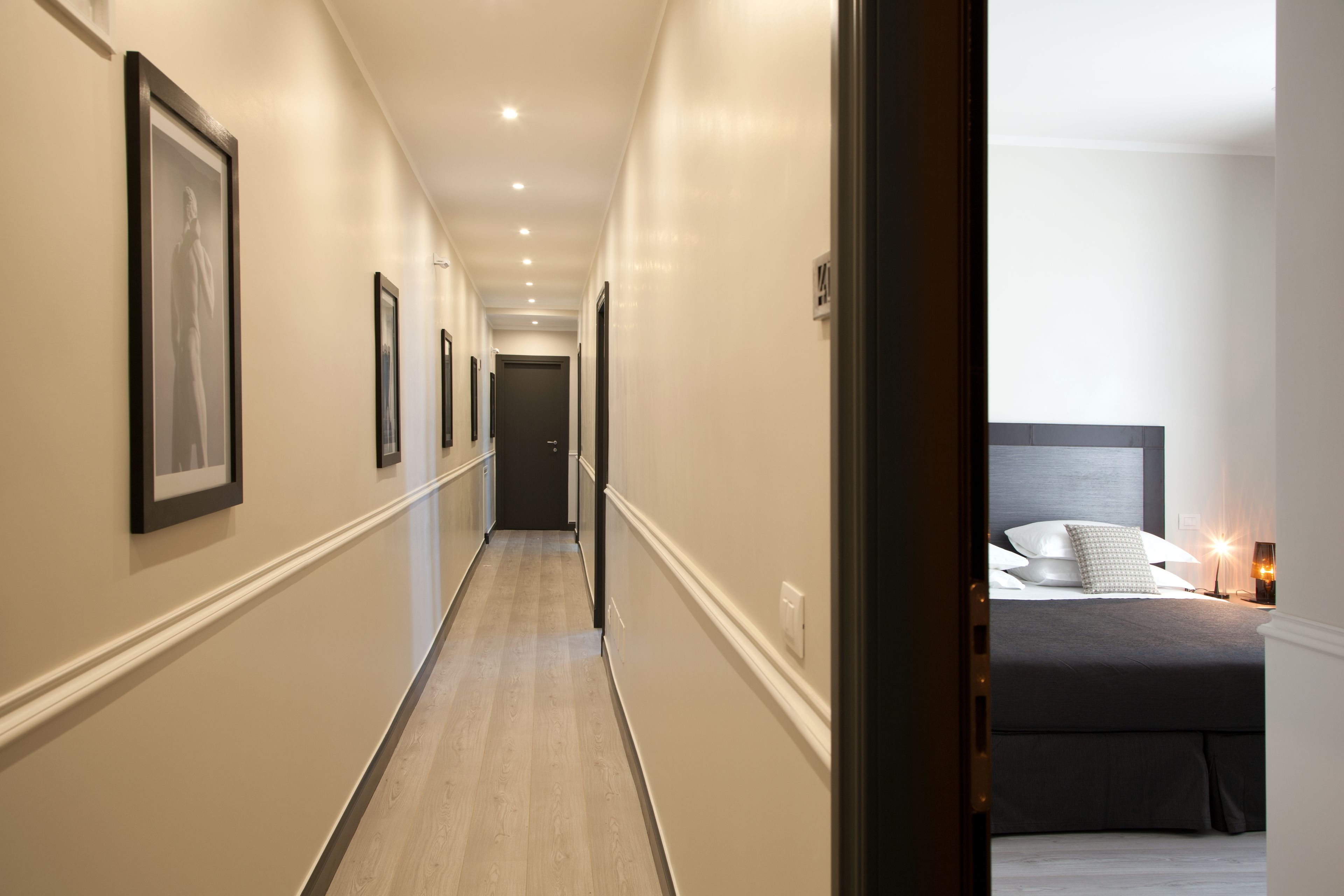 A hotel hallway with framed artwork on the walls, a bed with a headboard, and a bedside table with a lamp.